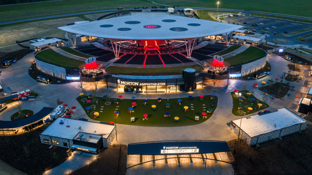 Aerial view of the Morton Amphitheater showcasing its modern architecture, surrounding landscaped area with colorful seating, and a spacious parking lot in the background.