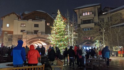 People gathered around a beautifully decorated Christmas tree in a festive outdoor setting at night, with buildings illuminated in the background.