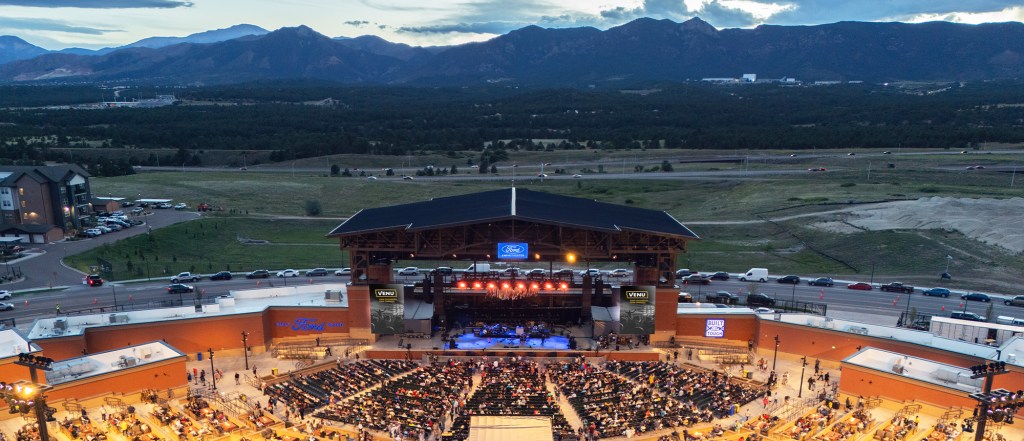 Aerial view of a concert venue with a large crowd seated in front of a stage, surrounded by mountains and greenery in the background.