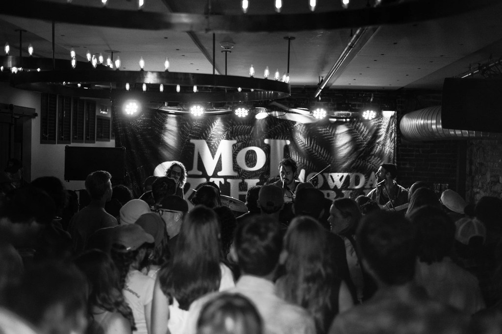A live music performance with a crowd in a dimly lit venue, featuring musicians on stage and a backdrop with the text 'MOL SHOWDA'.