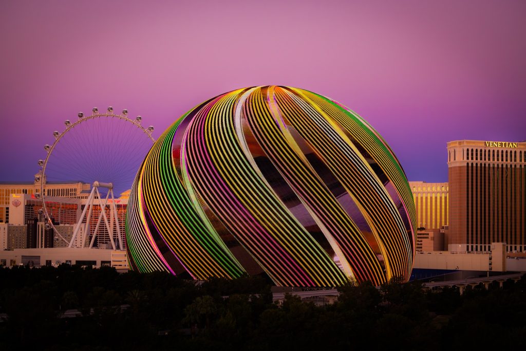 A colorful spherical structure illuminated at dusk, with a Ferris wheel and the Venetian hotel in the background against a purple sky.