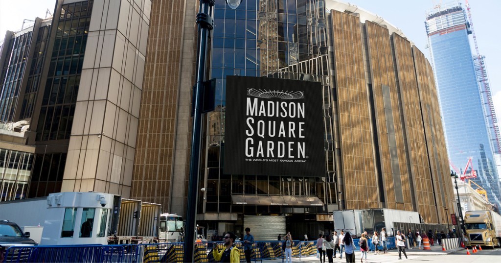 Exterior of Madison Square Garden with a large black sign displaying its name, surrounded by people and city buildings.