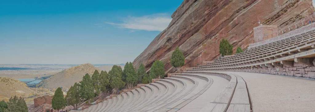 A panoramic view of the Red Rocks Amphitheatre, showcasing its unique natural stone backdrop and wooden seating arranged in a circular pattern, against a bright blue sky.