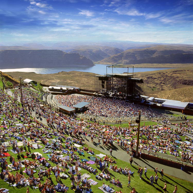 Aerial view of a large outdoor concert with a crowd gathered on grassy hills, with a lake and mountains in the background.